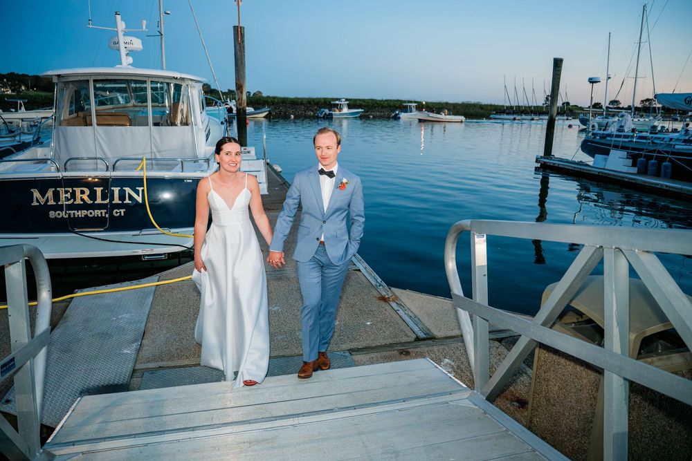 Couple walking along a marina dock with boats and blue water in the background at sunset.