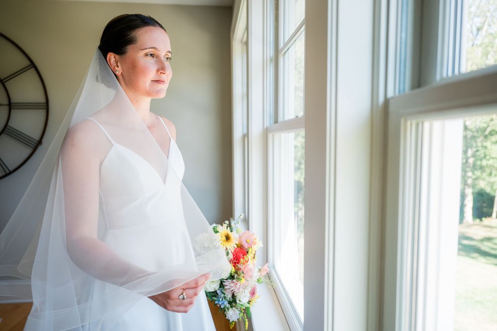 A bride stands in elegant white dress by large windows in a bright sunlit room with natural lighting.