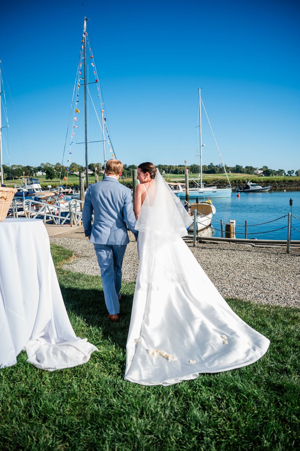 A wedding couple stands on grass near a marina with sailboats and blue water in the background.
