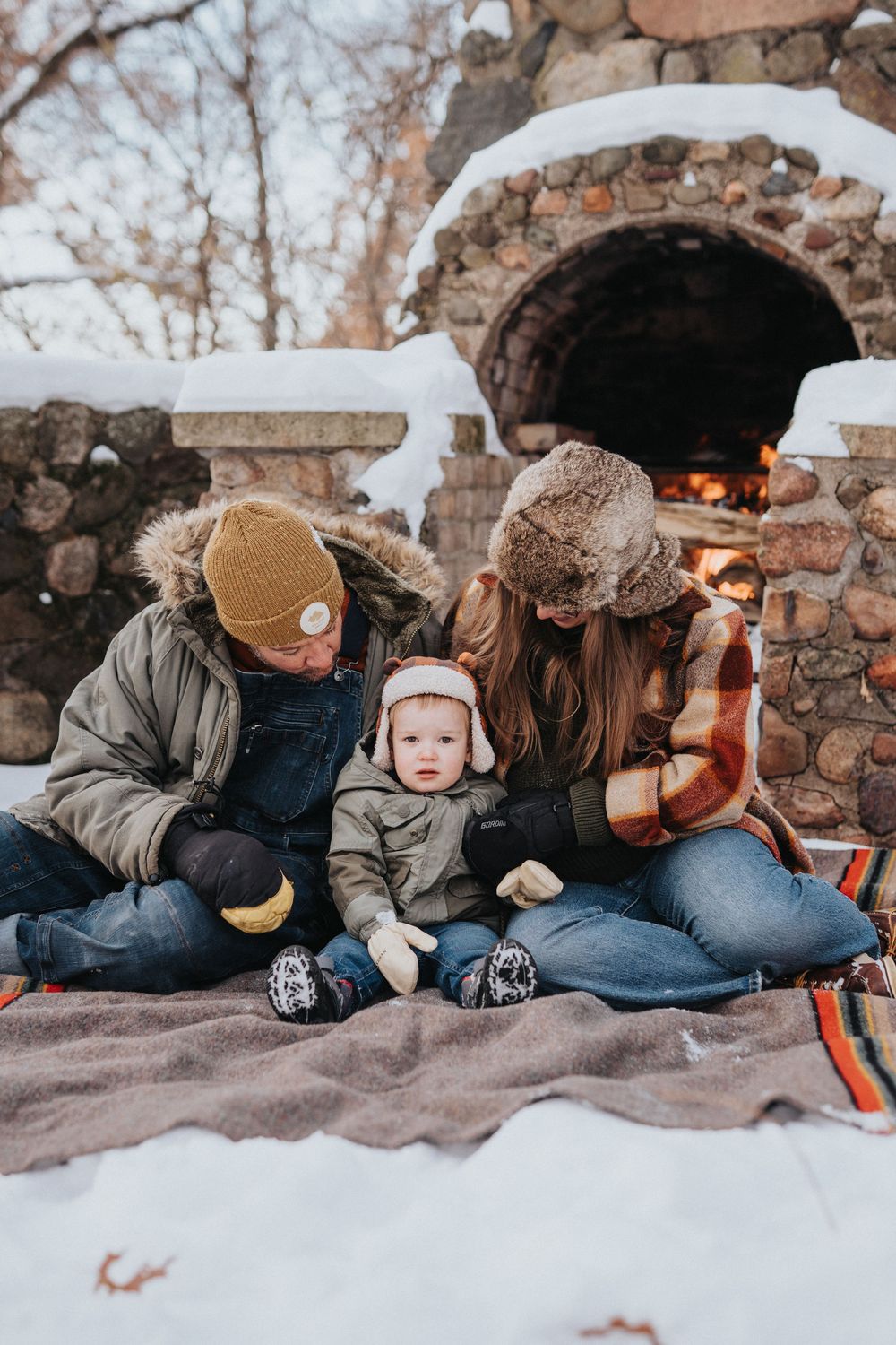 A family sits together on a blanket in the snow near a stone fireplace during a cozy winter outing.