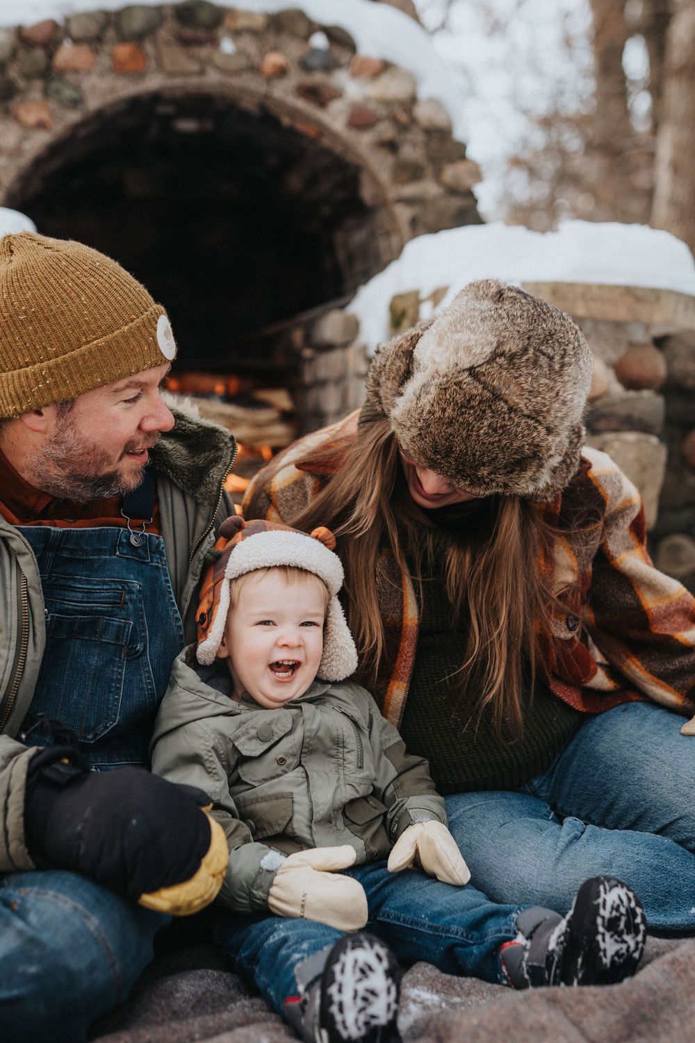 A family in winter clothing shares joyful moments together while sitting on snowy ground near a stone wall.