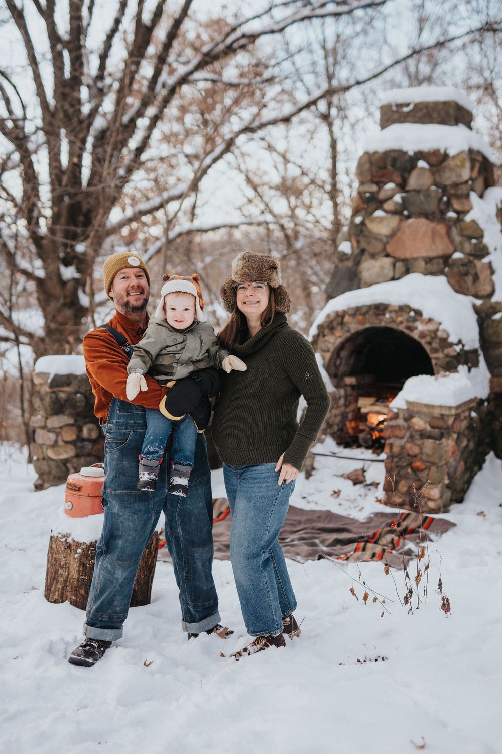 Winter family portrait session with casual poses in front of snow-covered trees and stone wall.