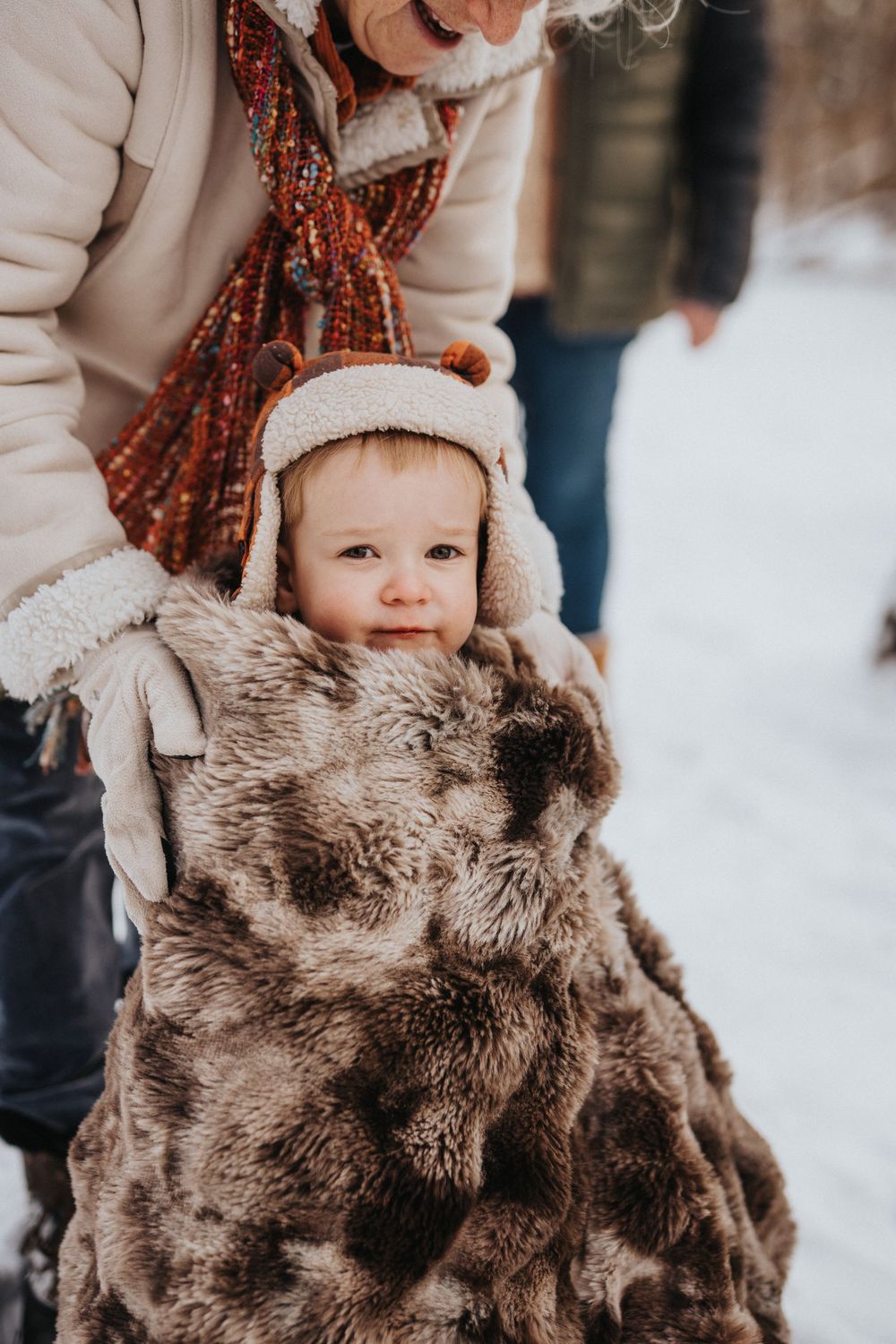 A small child in a winter coat holds a brown furry teddy bear in a snowy outdoor setting.