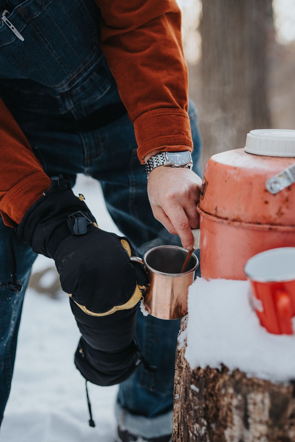 Person wearing orange jacket and jeans pours hot beverage from camping mug in snowy winter setting.