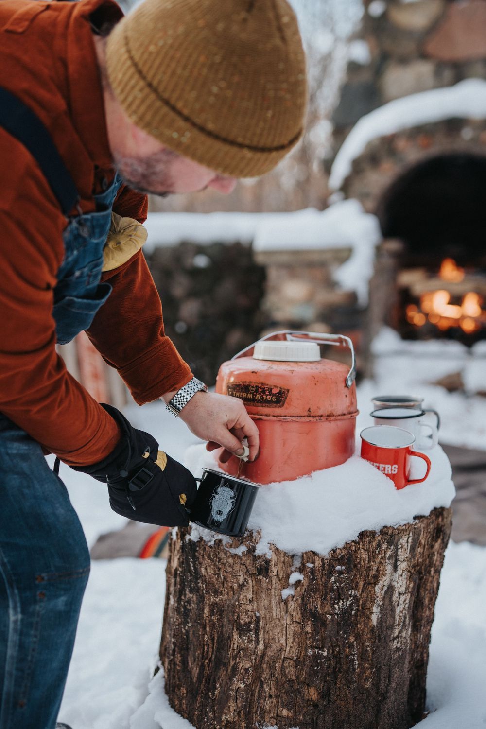 Person in orange jacket pours a drink from a thermos on a snowy tree stump with vintage camping gear.