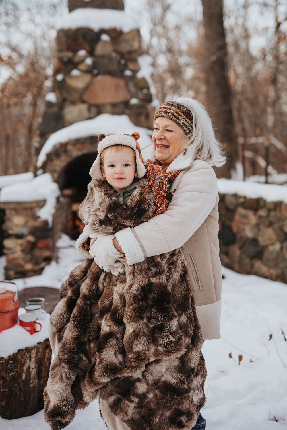 A cozy winter scene with two people in warm clothing hugging and smiling in front of a stone fireplace in the snow.