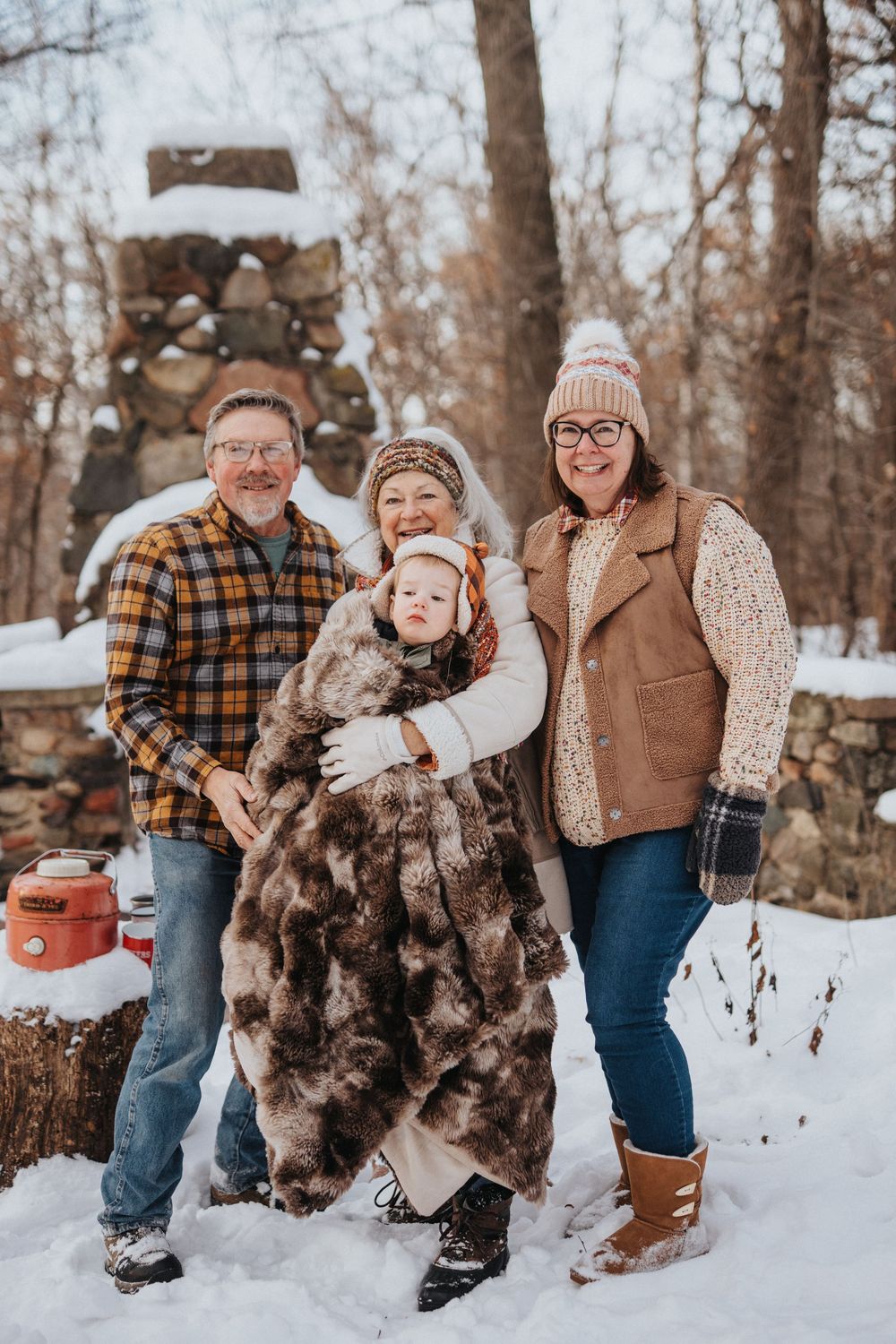 A family group poses together in winter attire against a snowy woodland backdrop with a stone structure behind them.