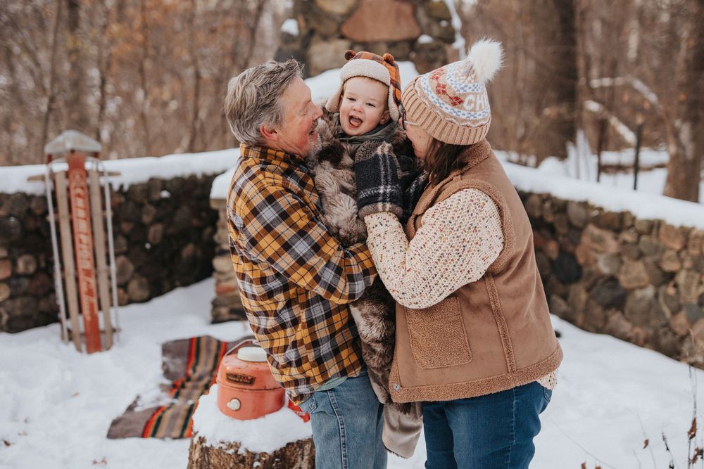 Family wearing cozy winter outfits embraces while enjoying a snowy outdoor photoshoot in a rustic woodland setting.