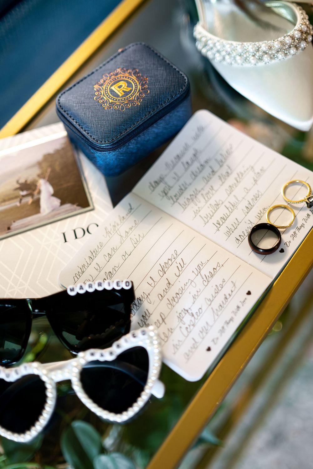 Wedding flat lay with rings, vows, sunglasses, jewelry box, and pearl accessories on a gold tray.