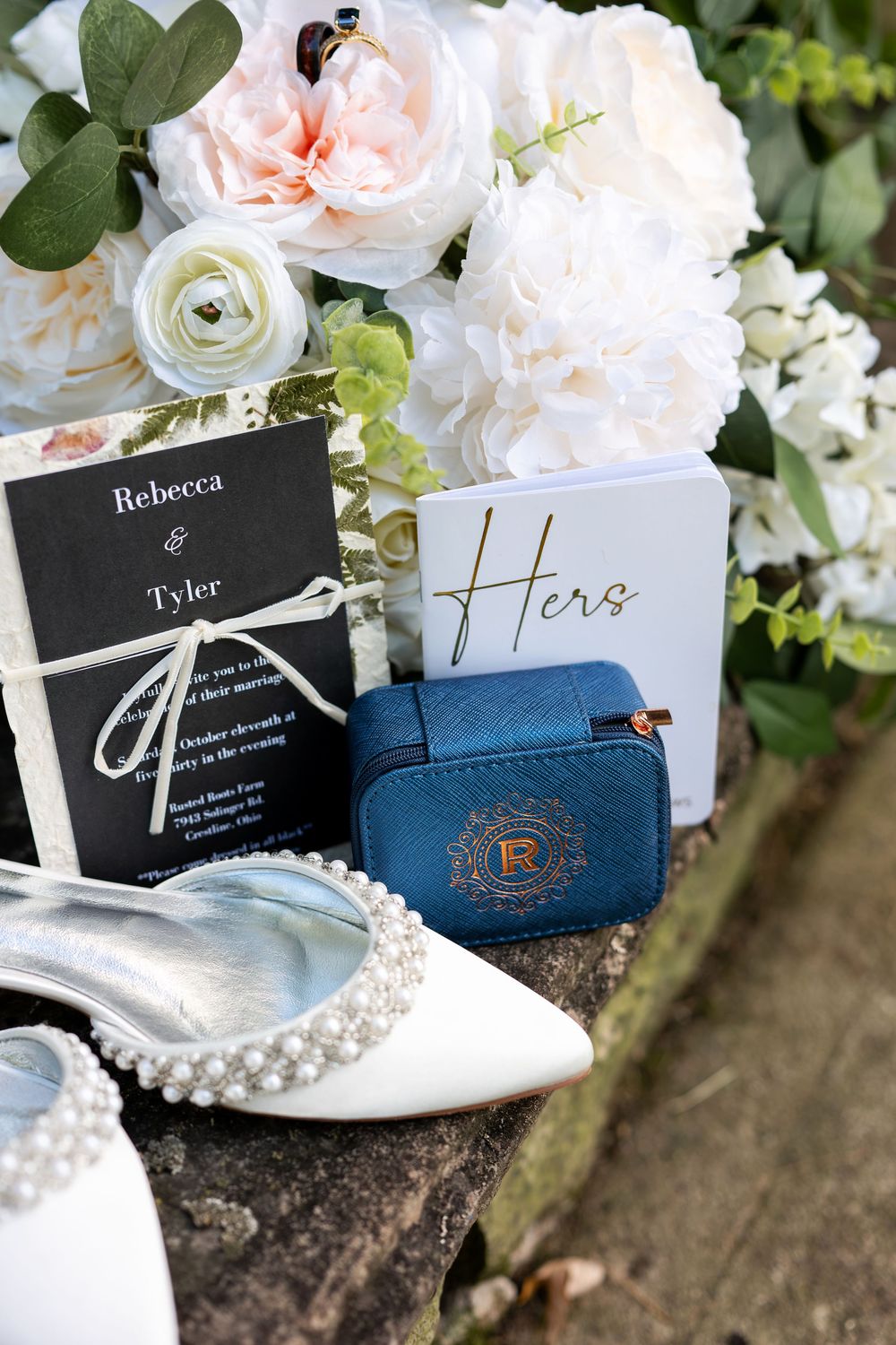 Elegant bridal flat lay featuring pearl shoes, Rebecca and Tyler wedding invitation, blue ring box, and white roses.
