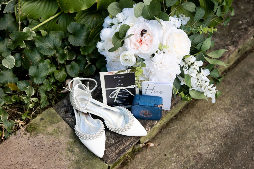Bridal detail flat lay with pearl heels, wedding invitation, blue ring box, and white floral bouquet on stone.