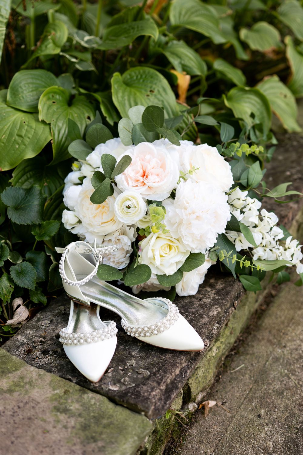 White bridal heels with pearl details rest beside a elegant bouquet of white and blush roses on stone steps.