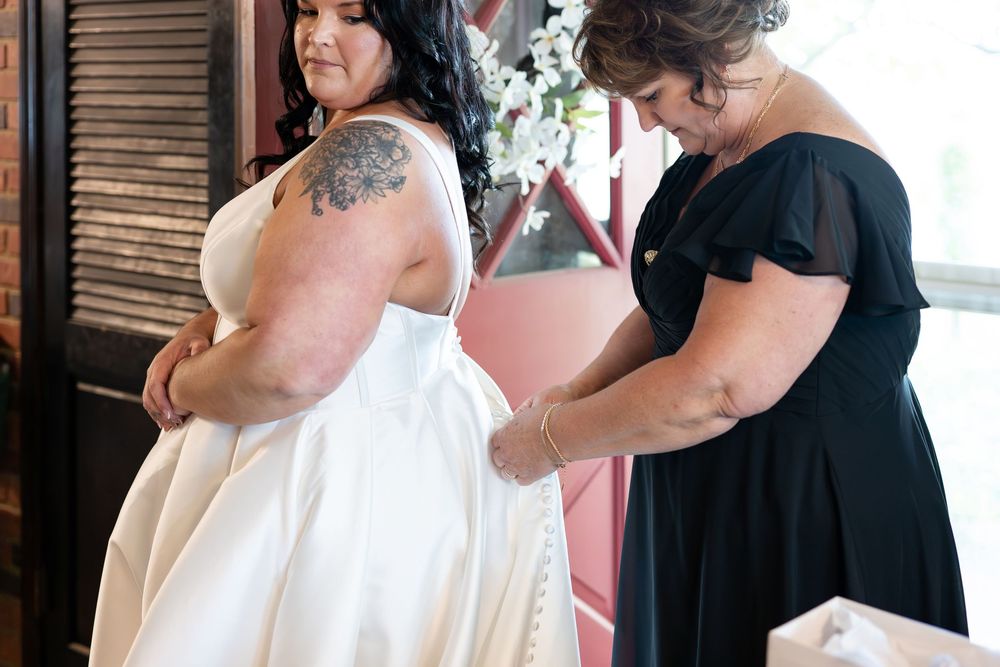 Bride in white wedding dress being helped with buttons by woman in black dress indoors.