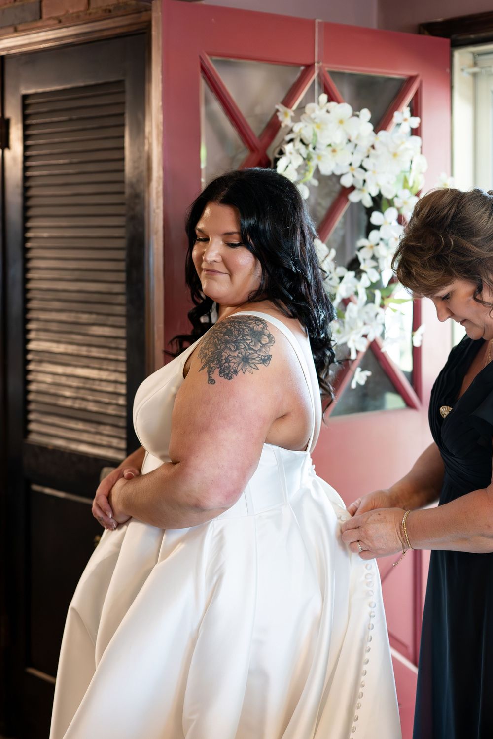 Smiling bride with tattoo in white gown being buttoned up by helper near floral decoration.