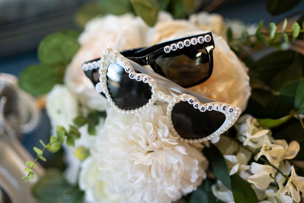 Two decorative sunglasses reading groom and bride rest on white wedding bouquet flowers.