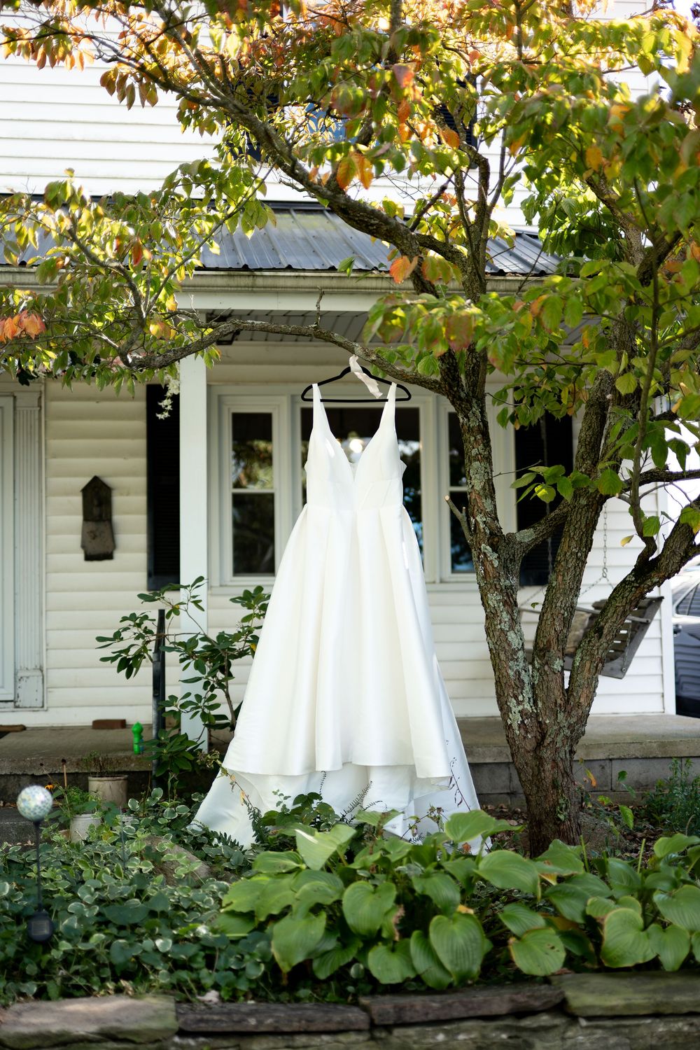 White wedding dress hanging on a tree branch outside a charming white farmhouse surrounded by lush greenery.