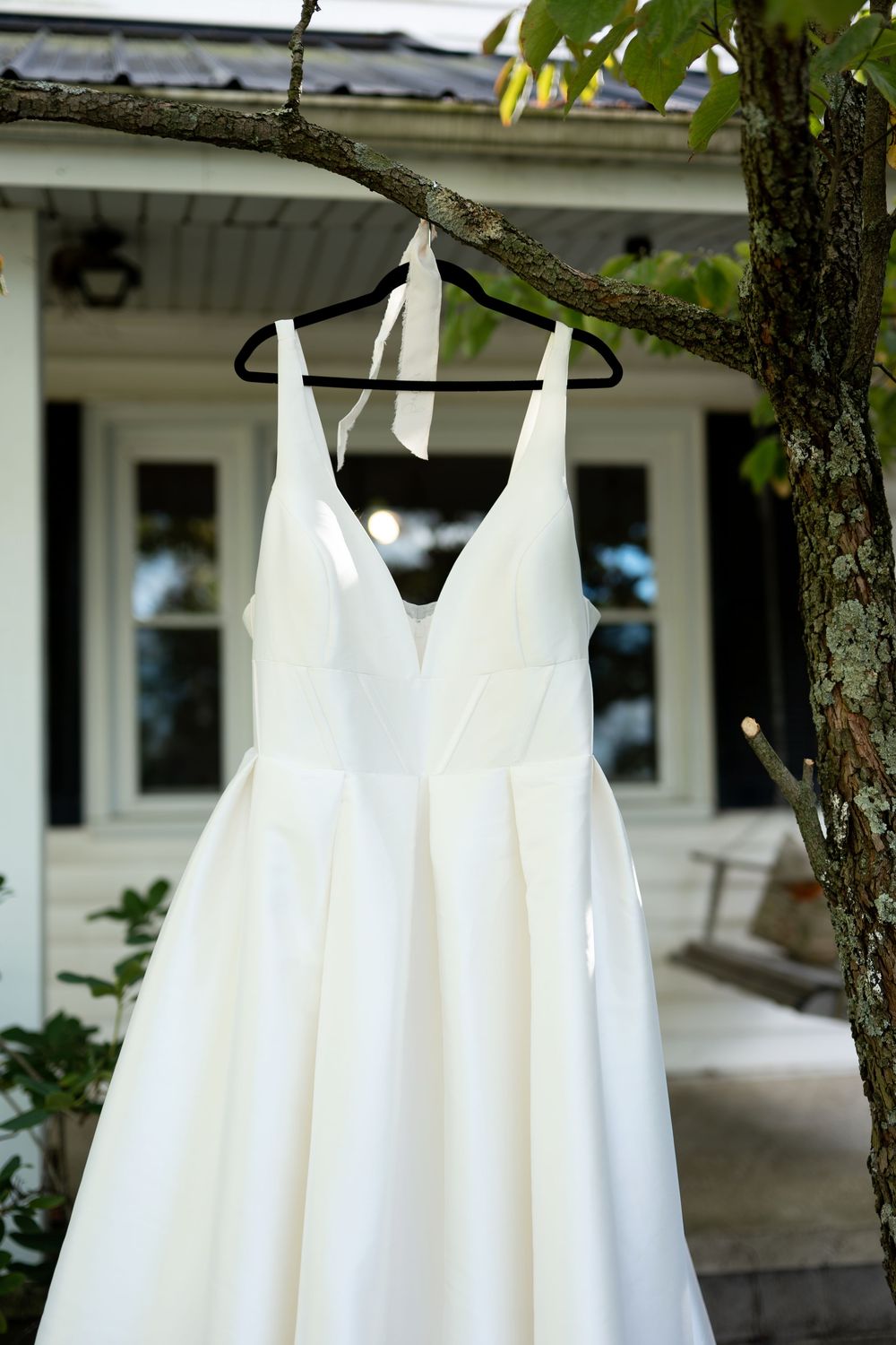 Elegant white V-neck wedding gown hanging on a tree branch outside a white house.