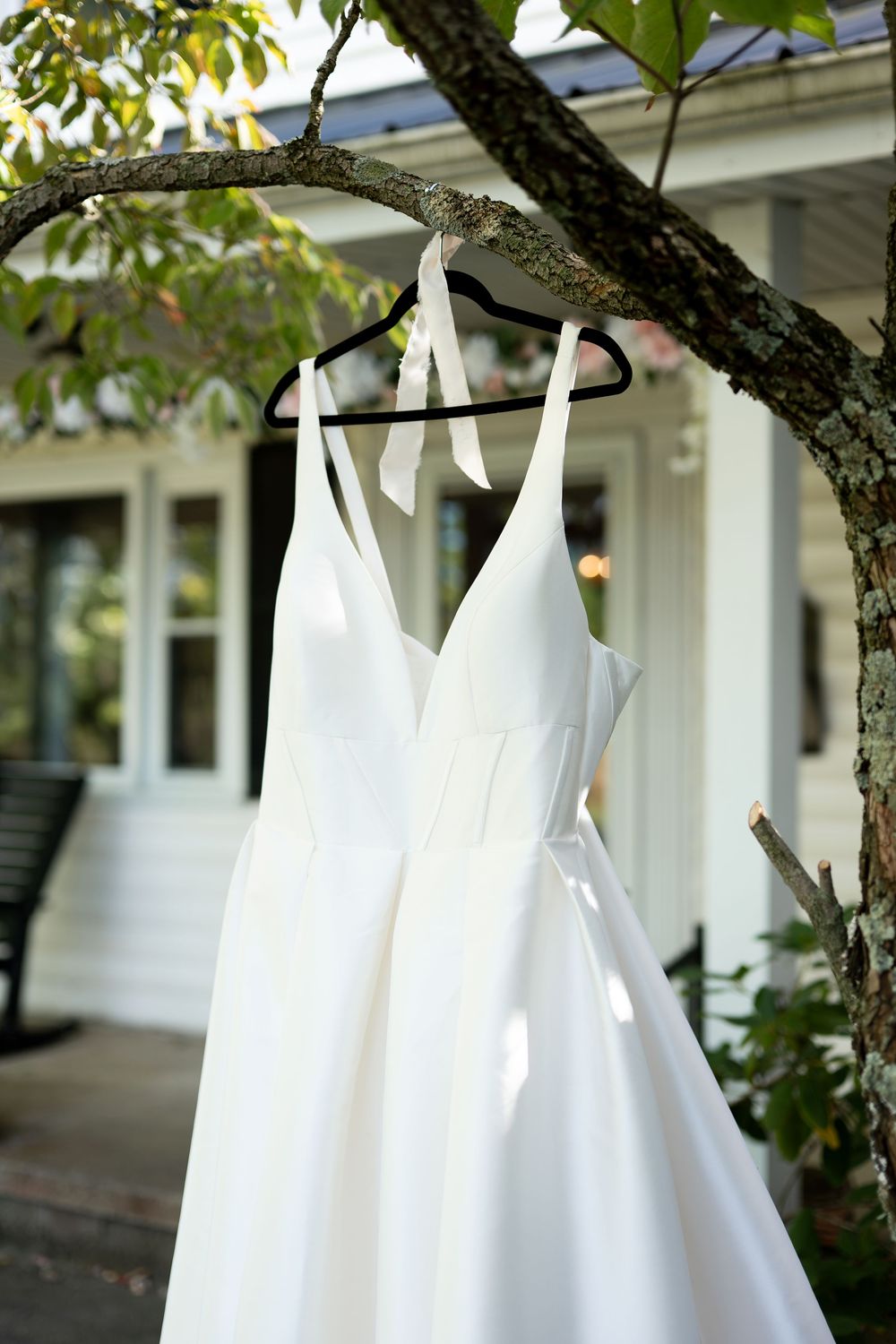 Elegant white bridal gown with V-neck and ribbon straps hanging on a black hanger from a tree branch.