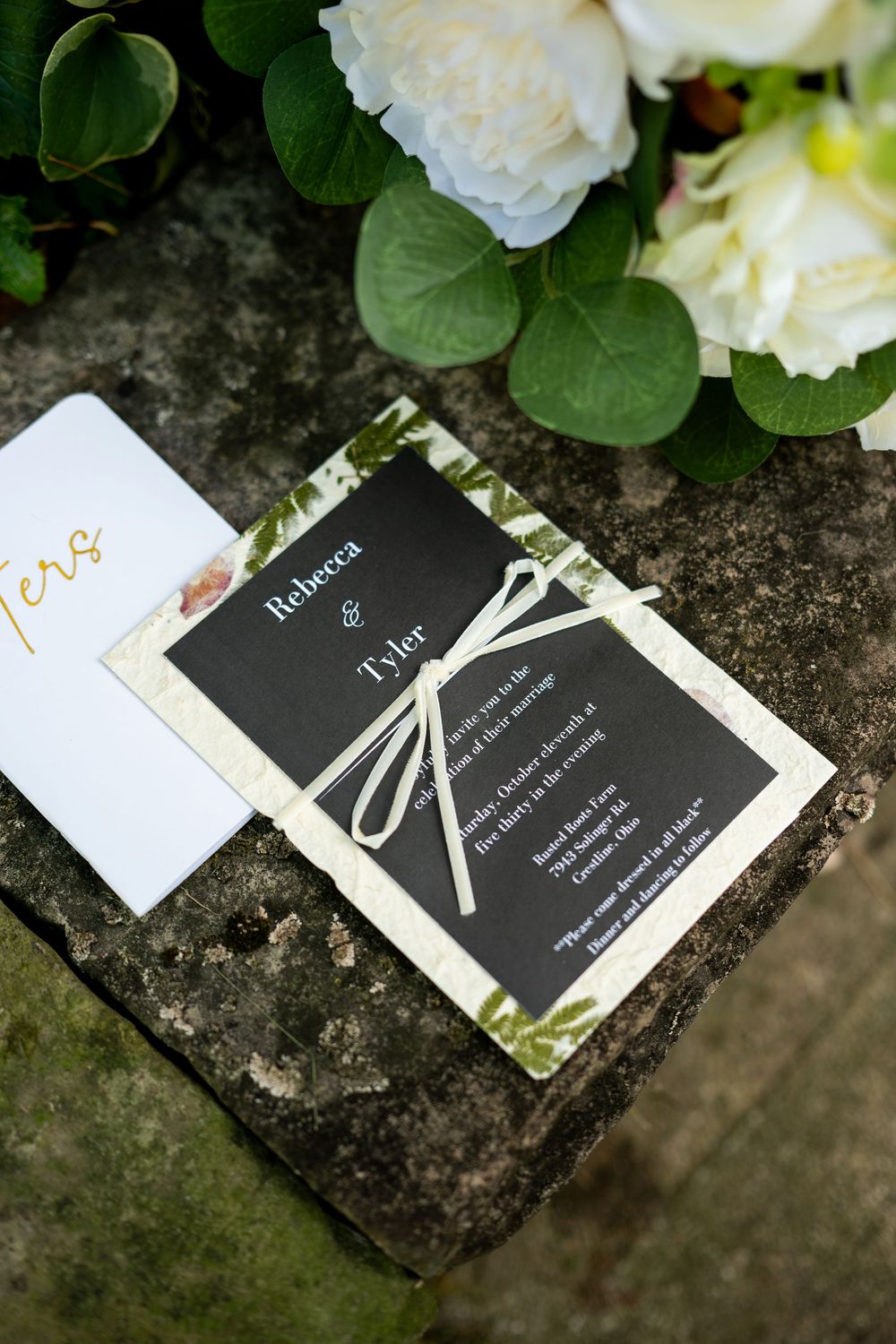 Black and white wedding invitation for Rebecca and Tyler tied with ribbon resting on stone beside white roses.