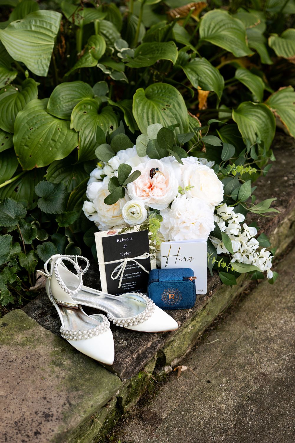 White pearl ankle-strap bridal shoes displayed with bouquet, wedding invitation, and blue ring box on stone steps.