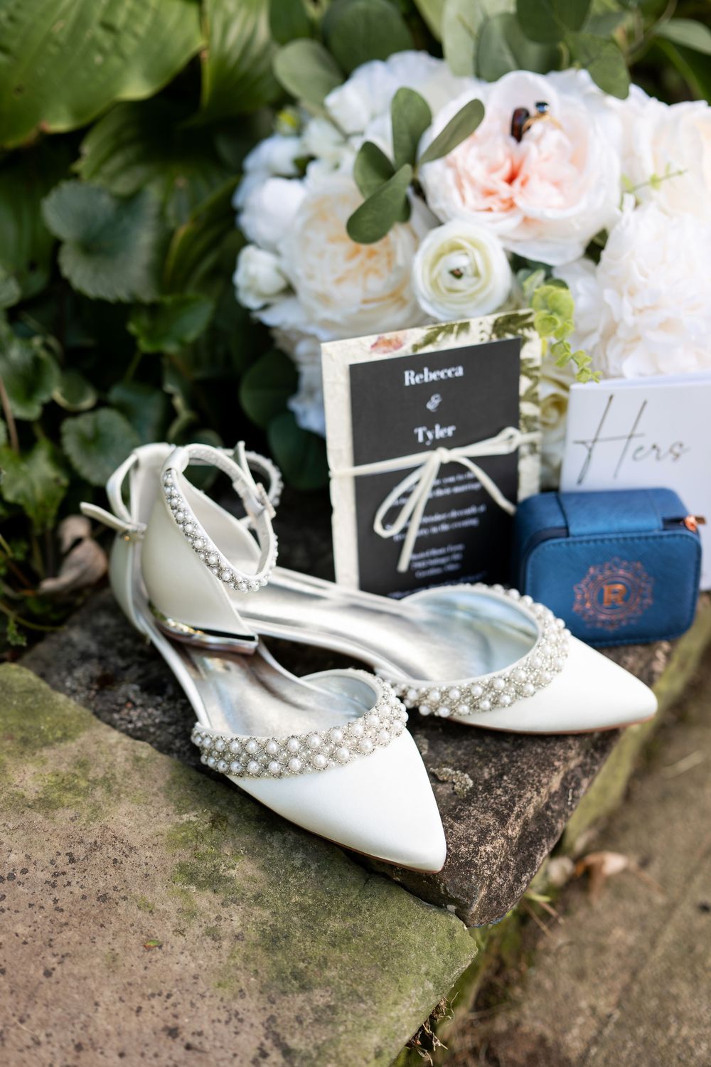 Pearl-embellished white bridal heels displayed with wedding invitation, blue ring box, and floral bouquet on stone.