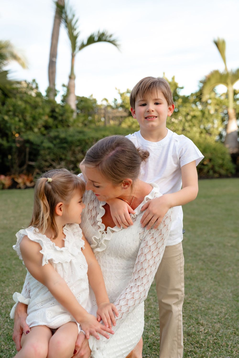 A playful family photoshoot in white outfits on a grassy lawn with palm trees in the background.