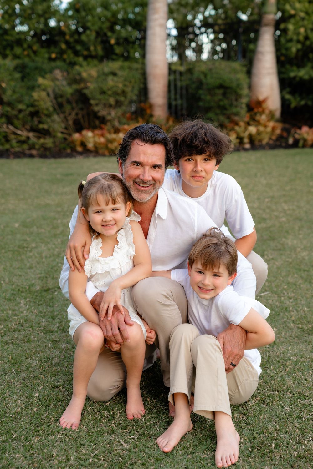 Family members in coordinated white and beige outfits sitting together on grass during a photo session.