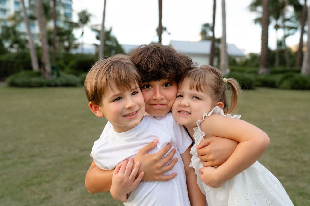 A group embraces warmly outdoors in a grassy area with palm trees visible in the background.