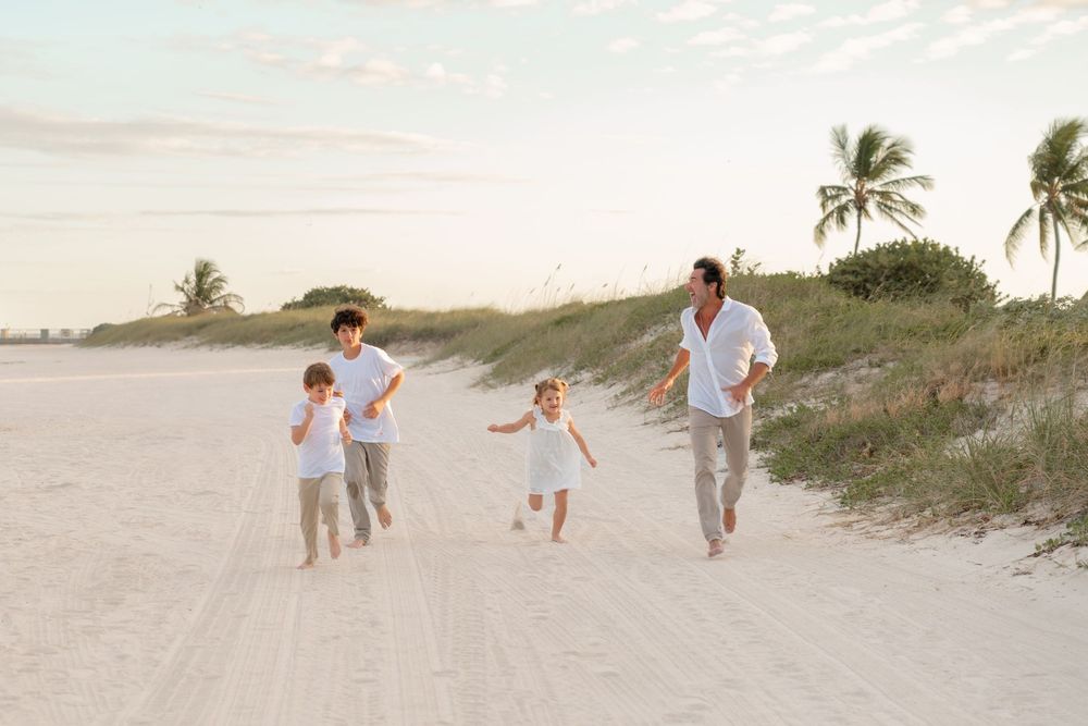 A family walks together along a sandy beach path with palm trees and dunes at sunset.
