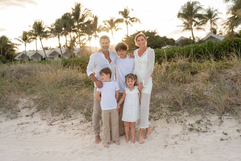 Family wearing white and beige clothing poses together on a sandy beach at sunset with palm trees.