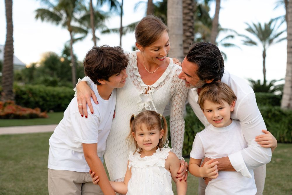 A family in coordinated white and beige outfits embraces while posing for a portrait in a tropical garden setting.