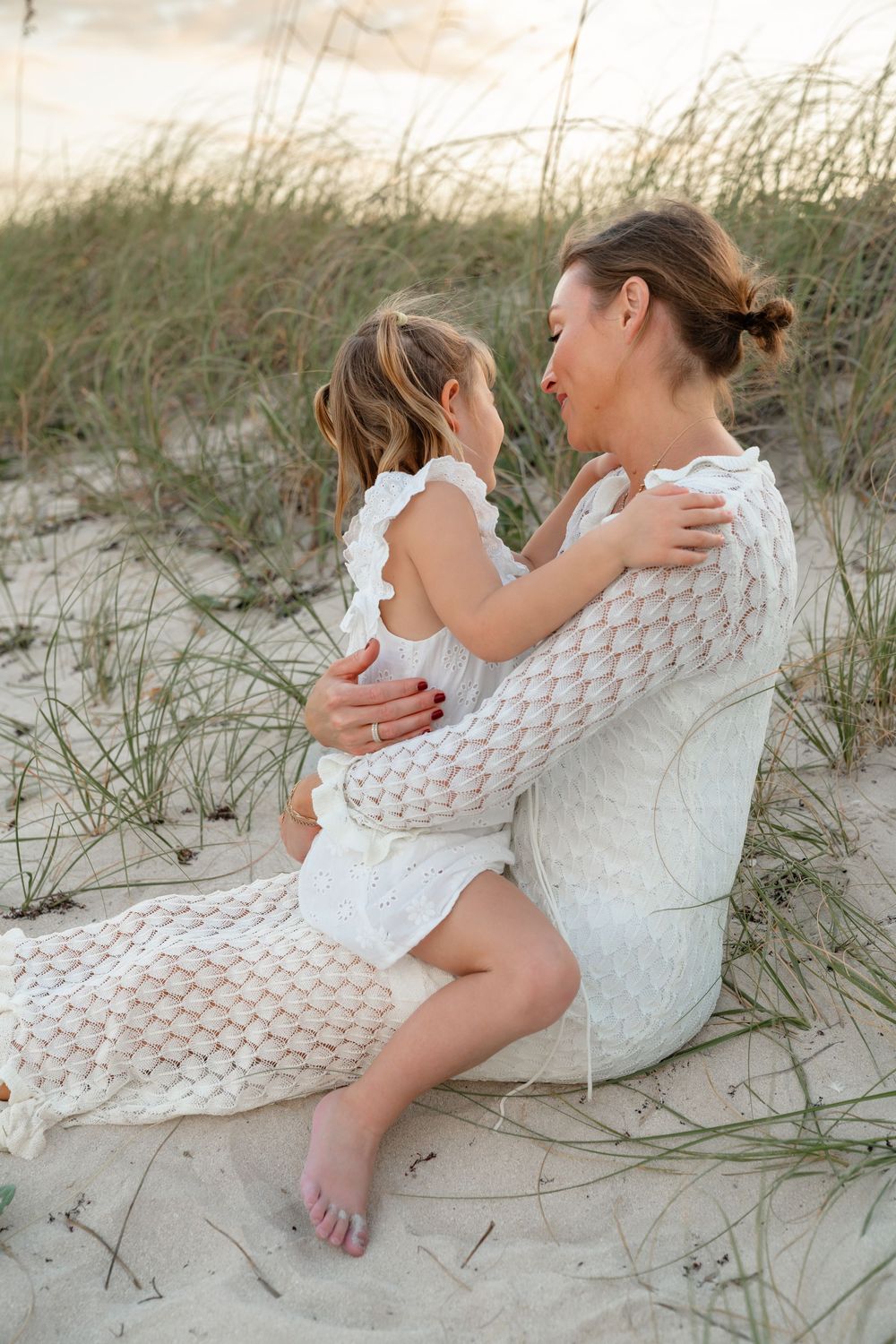 Mother and child share tender embrace while sitting on sandy beach dunes at sunset.