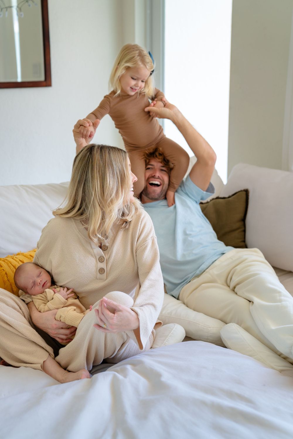 Family enjoying playful moments together on white bedding while wearing cozy beige outfits.