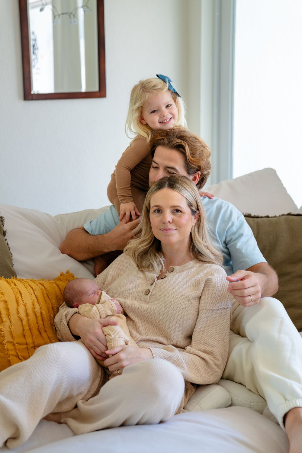 Parent and baby in matching beige outfits cuddle on a cozy grey couch with yellow accent pillows.