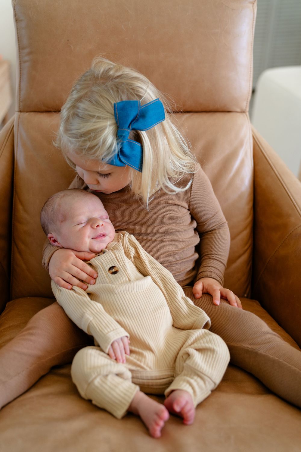 Baby wearing cream colored outfit rests peacefully on tan leather couch.