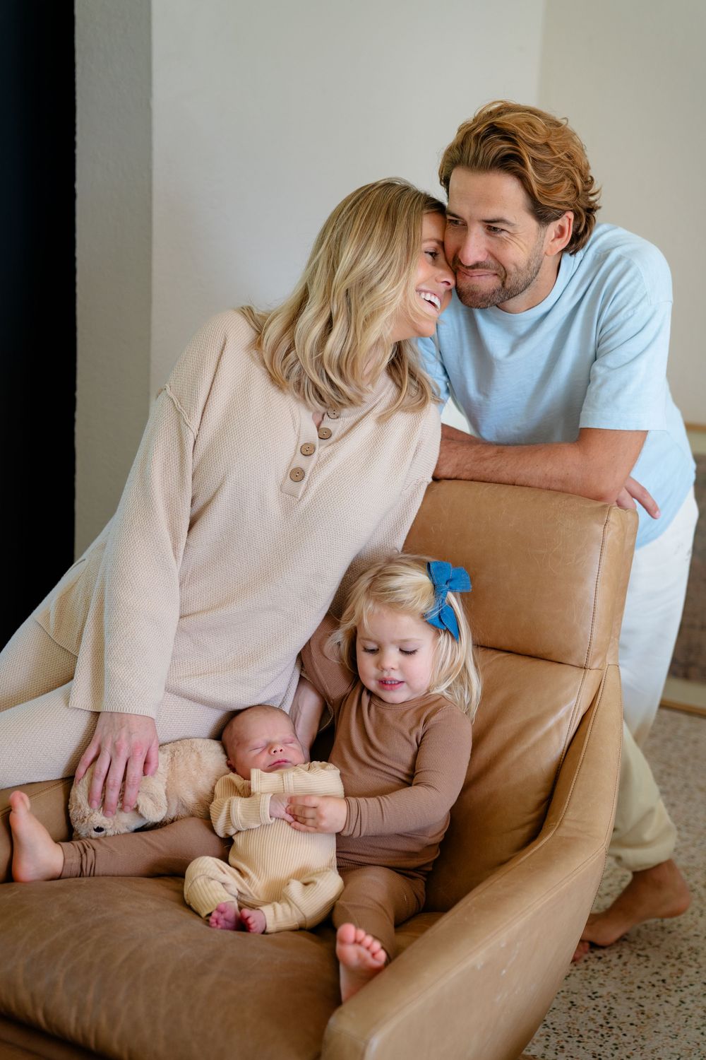 A family shares a tender moment together on a brown leather chair with their baby and young child.