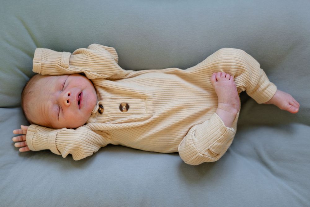 Sleeping baby in cream ribbed onesie lies stretched out on grey cushions.