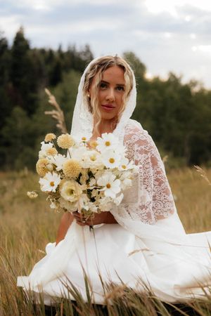 A bride in lace sitting in tall grass holding a white and yellow bouquet.