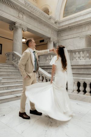 A couple in wedding attire twirls and dances on an elegant marble staircase in a grand building.