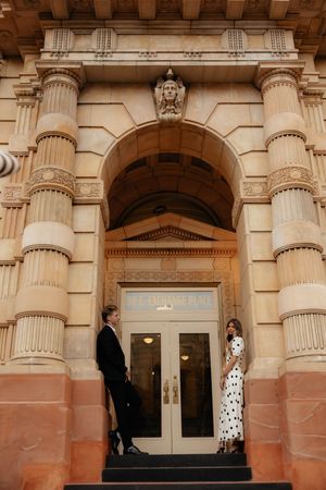 Elegant couple poses beneath grand classical columns of historic building entrance.
