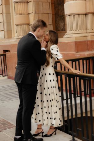 Couple in black suit and polka dot dress pose playfully on ornate building steps.