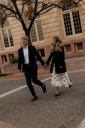 Couple in formal wear dance and play among autumn leaves outside historic building.