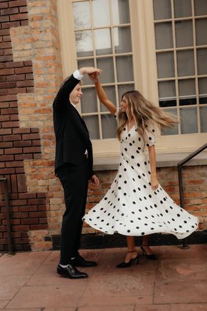 Woman in flowing polka dot dress twirls while dancing with partner by brick building.