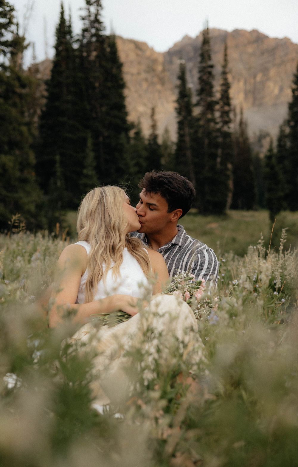 A romantic moment shared in a wildflower meadow with mountain peaks and pine trees in the distance.