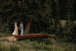 A couple walks along a mountain trail surrounded by evergreen forest and distant peaks.
