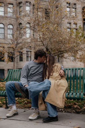 A couple shares a playful moment on a green park bench with autumn leaves and a historic building in the background.