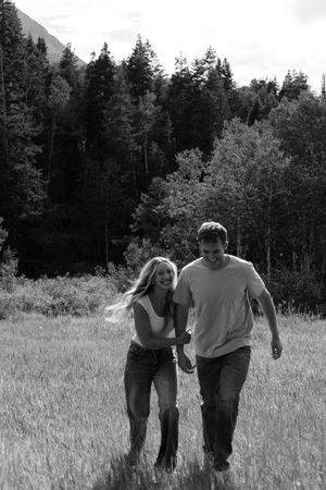 A couple walks through a sunlit meadow holding hands in black and white.