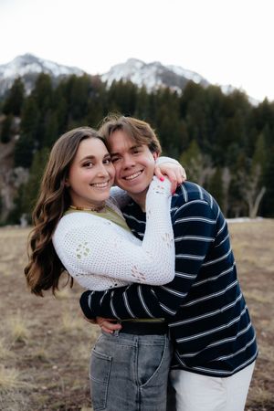 Two young adults, a man and a woman, embrace outdoors in a scenic mountain landscape.