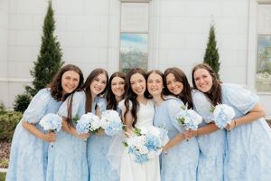 Bride in white gown with six bridesmaids in light blue dresses holding blue hydrangea bouquets outdoors.