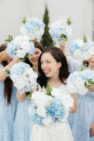Bride in white dress with bridesmaids in blue, holding matching blue hydrangea and white rose bouquets outdoors.